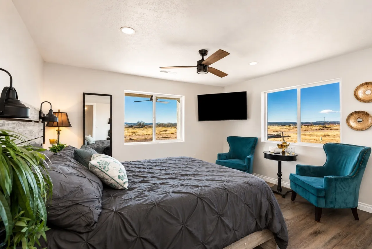 Garden Suite primary bedroom with a king bed, wall-mounted TV, teal velvet wingback chairs, and desert views out two windows.